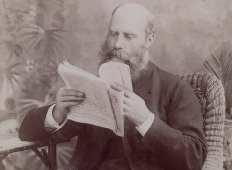 A formal portrait of Bishop Sydney Linton. He is seated in a cane chair reading some papers